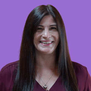 Smiling woman with long brown hair wearing a maroon top and heart necklace, set against a bright purple background.