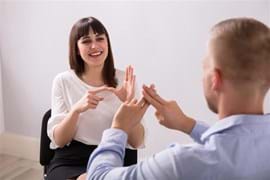 two people sitting opposite one another, the man has his back to the camera wearing a blue shirt, the lady is facing the camera wearing a white blouse and black shirt. They are communicating using signing and their hands are animated..