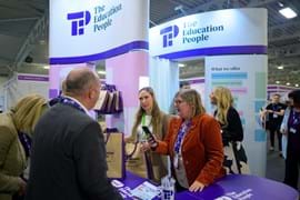 A group of smiling people engage in conversation at The Education People booth at a conference. The booth displays services like professional development.