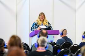 A woman with long blonde hair speaks at a podium with a purple cover. She is smiling and holding papers. Audience members wear headphones