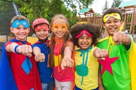 Five children (boys and girls) all wearing bright coloured outfits looking like super hero's, wearing capes and all with their left arms out in front of them like a 'superman' pose