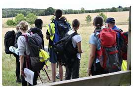 Mixed group of students, boys and girls, all in walking gear and carrying heavy rucksacks, some holding maps, walking through a field, looks like they may have just climbed over a sty
