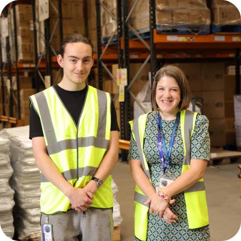 ALT Two people wearing reflective safety vests smile in a warehouse. Surrounding them are shelves with cardboard boxes. The atmosphere is friendly and professional.