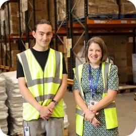ALT Two people wearing reflective safety vests smile in a warehouse. Surrounding them are shelves with cardboard boxes. The atmosphere is friendly and professional.
