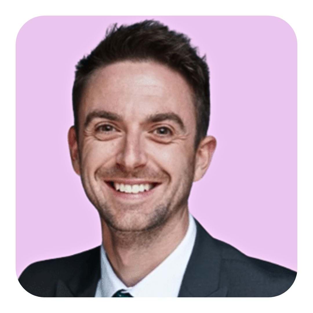 Smiling man in a suit and tie against a pink background. He has short dark hair and a friendly expression, conveying a professional and approachable tone.