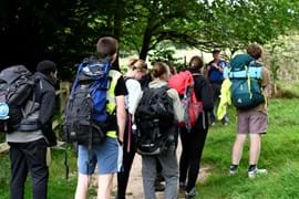 Group of Duke of Edinburgh Award students on an expedition, all with large rucksacks on their back, walking in fields with trees in the distance