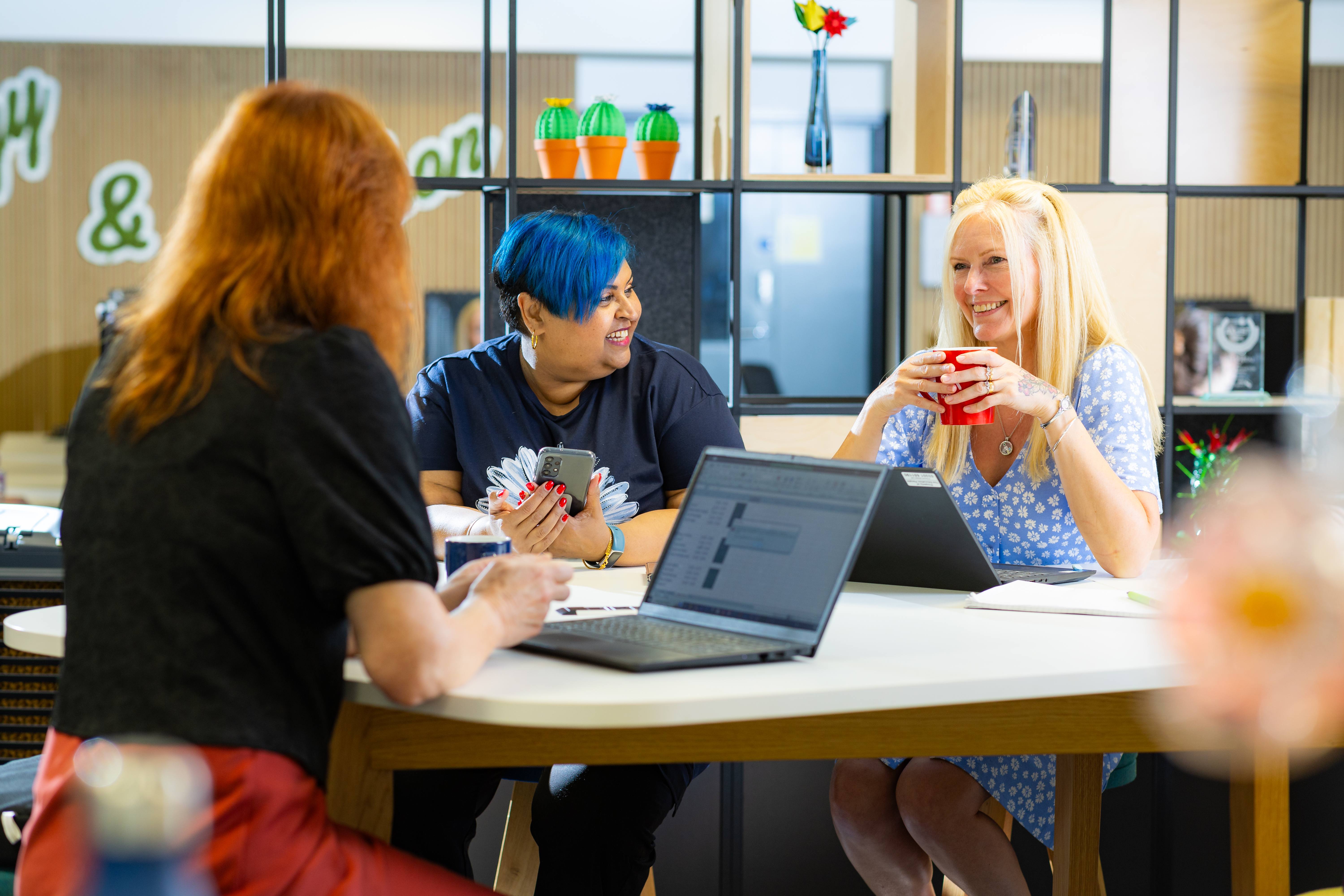 Three ladies sitting round a desk with laptops in front of them. One lady is holding a cup with both hands smiling, the other is smiling at her holding a mobile phone, the other lady is listening to the conversation.