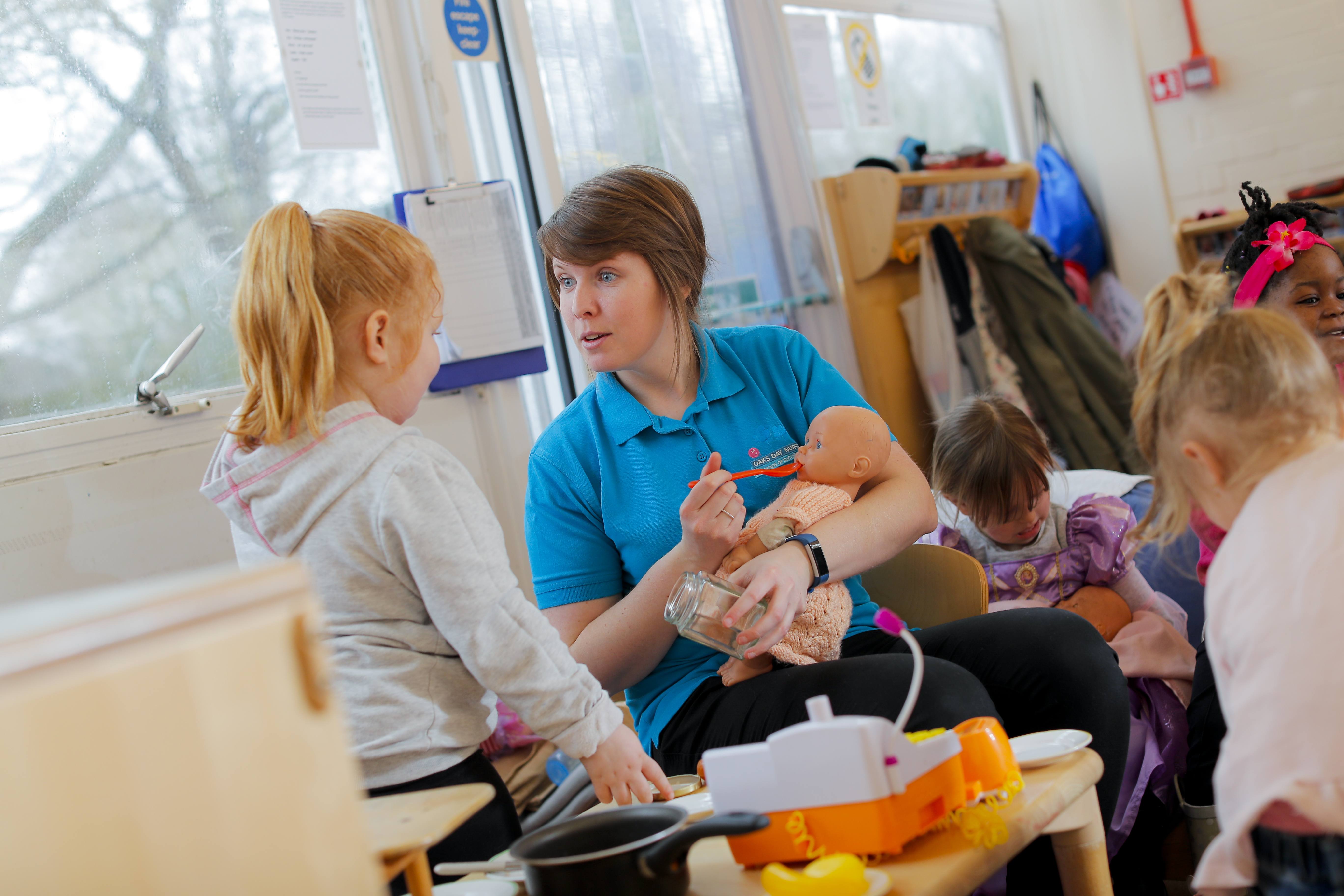 Nursery teacher sitting holding a dolly and pretending to feed her whilst talking to a child, other children are playing in the background