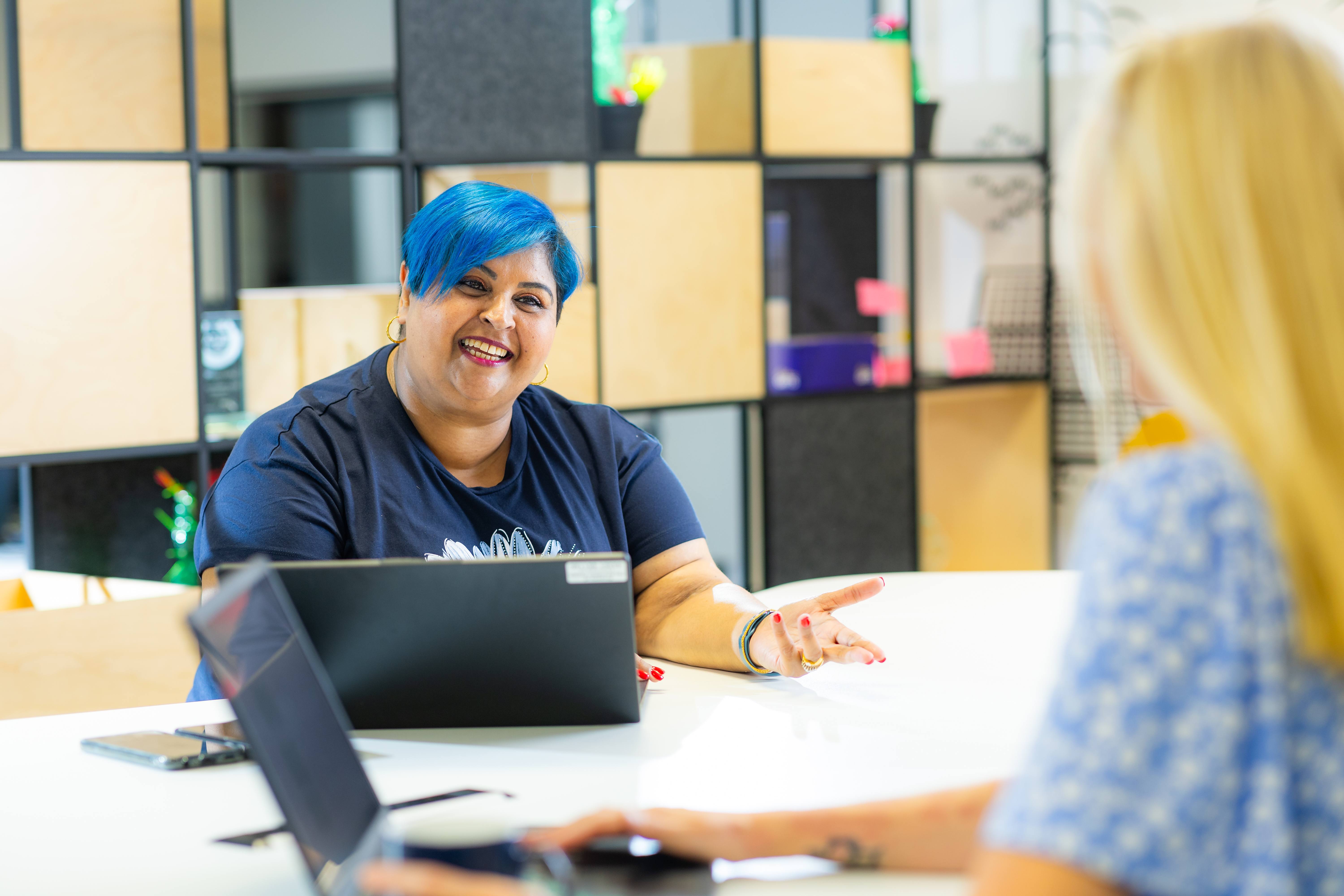 Two ladies sitting at a desk working on laptops, one lady is using her hands to express herself in conversation whilst the other listens