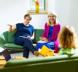 Three ladies sitting across from each other with notepads and pens having a conversation, one is using her hands to express herself, the other is holding a yellow book