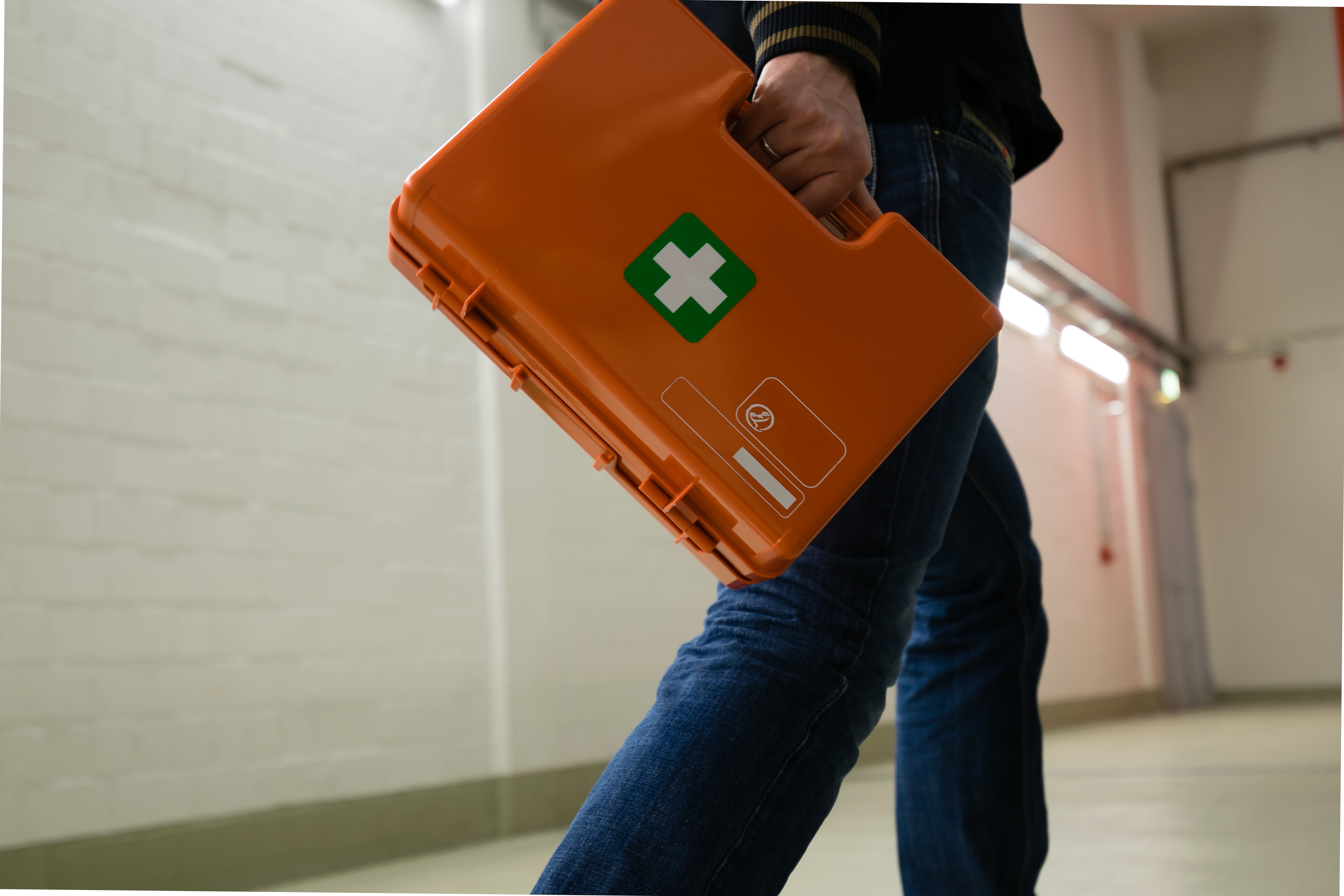 ALT - A person walks down a corridor holding an orange first aid kit with a white cross, wearing jeans.