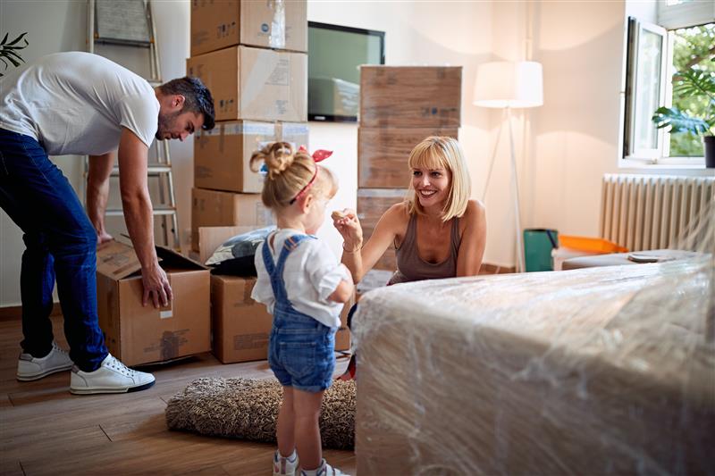 A family unpacks in a sunlit room. A mother smiles at a young daughter with a red bow, while the father organizes boxes.
