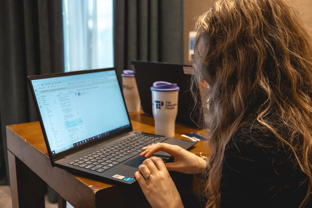 A woman with long curly hair sits at a wooden table, focused on her laptop with an open email.
