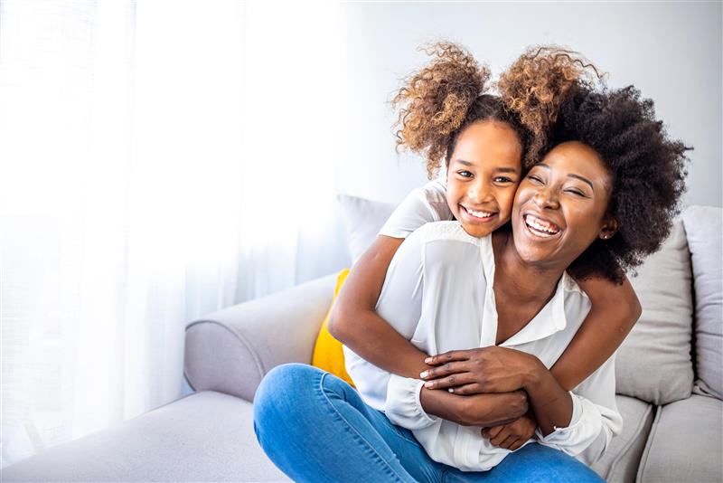 A joyful mother and daughter with curly hair embrace on a couch, smiling brightly.