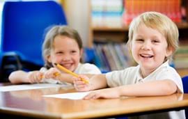 ALT - two children sitting at a desk holding pencils ready to write on the paper in front of them.