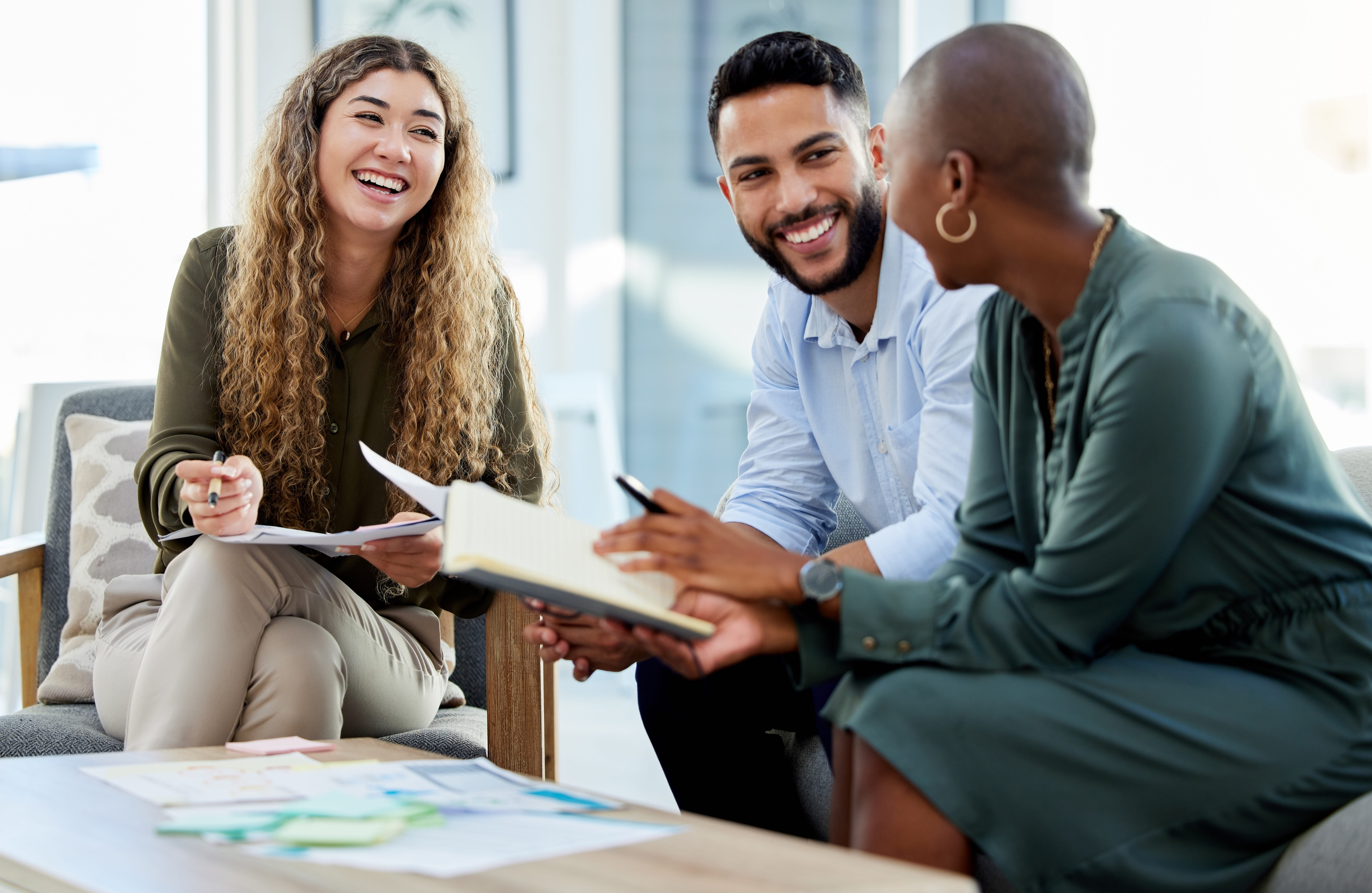 Three people sit in an office lounge, smiling and engaged in a discussion.