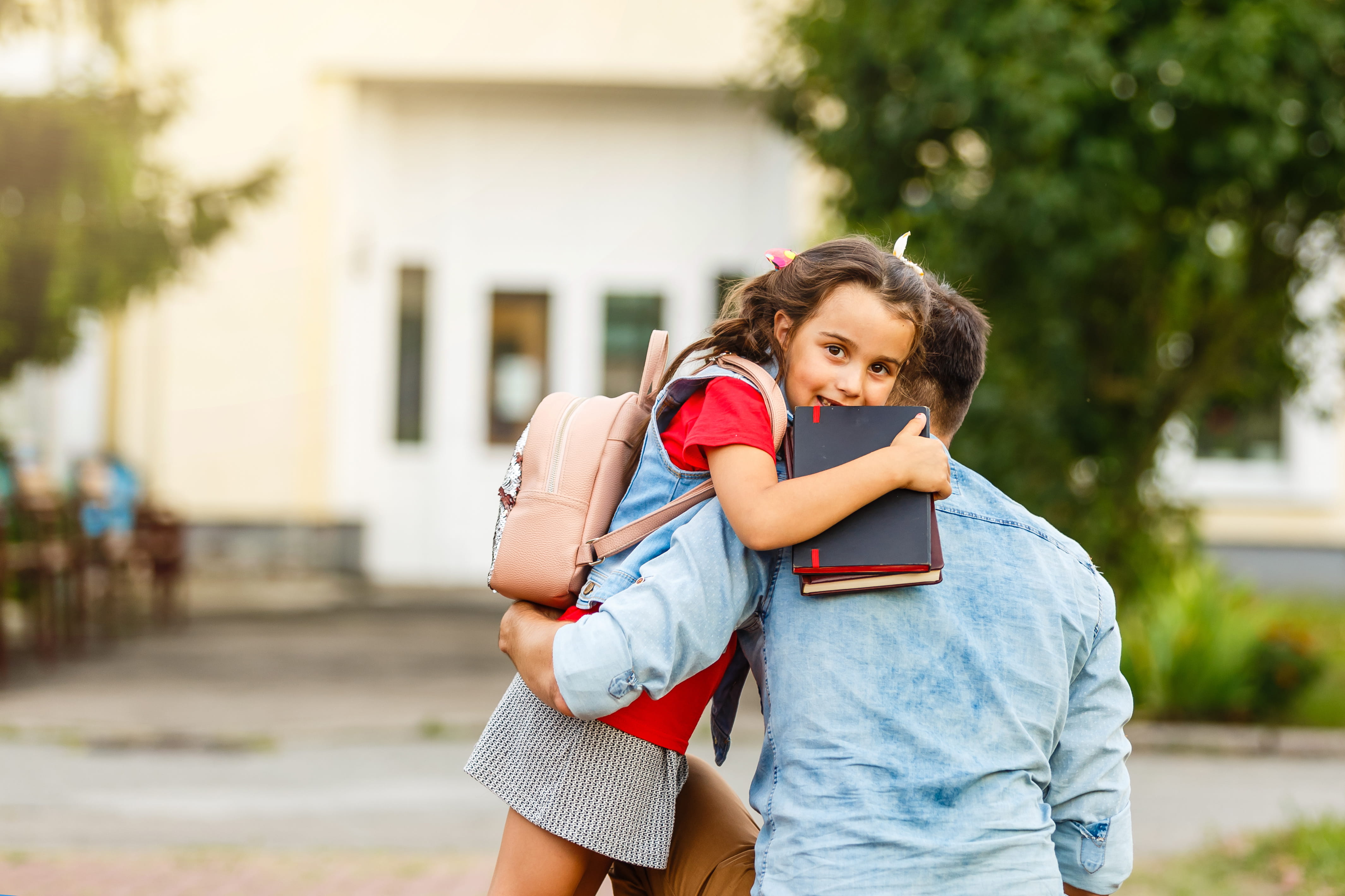 ALT - A young girl with a backpack is being hugged by a man outside a building.