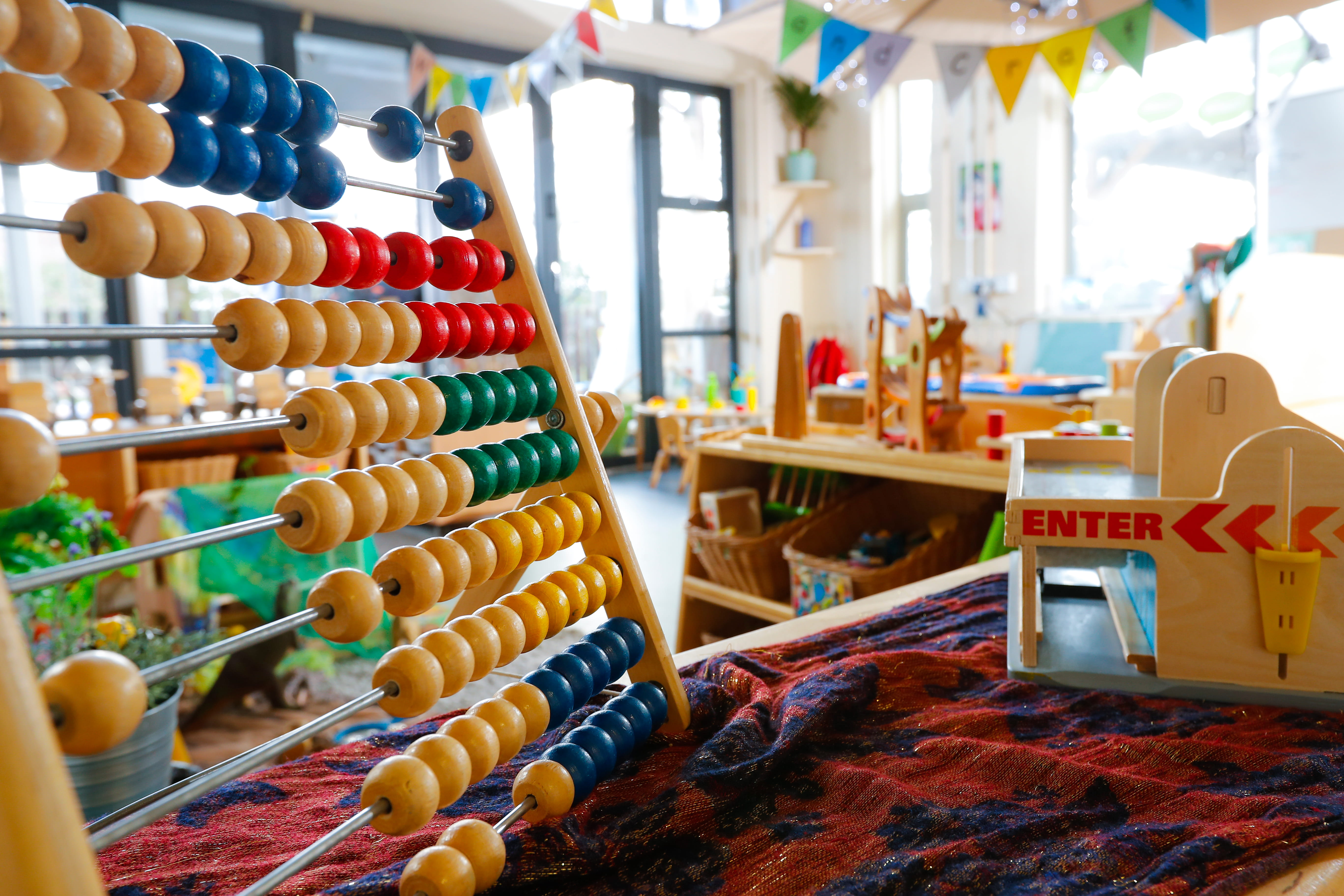 ALT - Wooden abacus with colourful beads sits in a bright, inviting classroom filled with toys and educational materials.