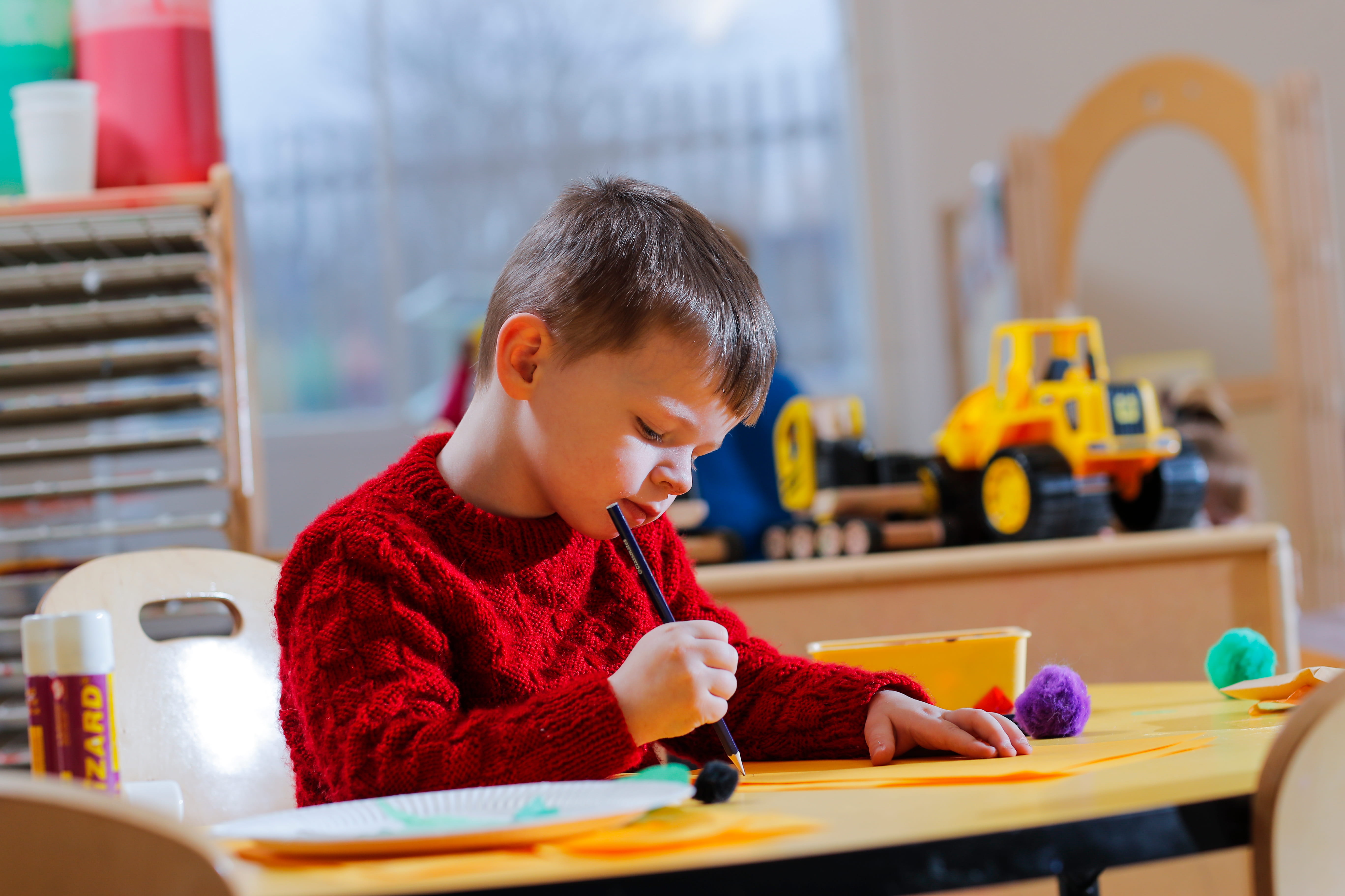 ALT - Young boy in a red sweater focused on painting at a classroom table. Bright toys and art supplies surround him.