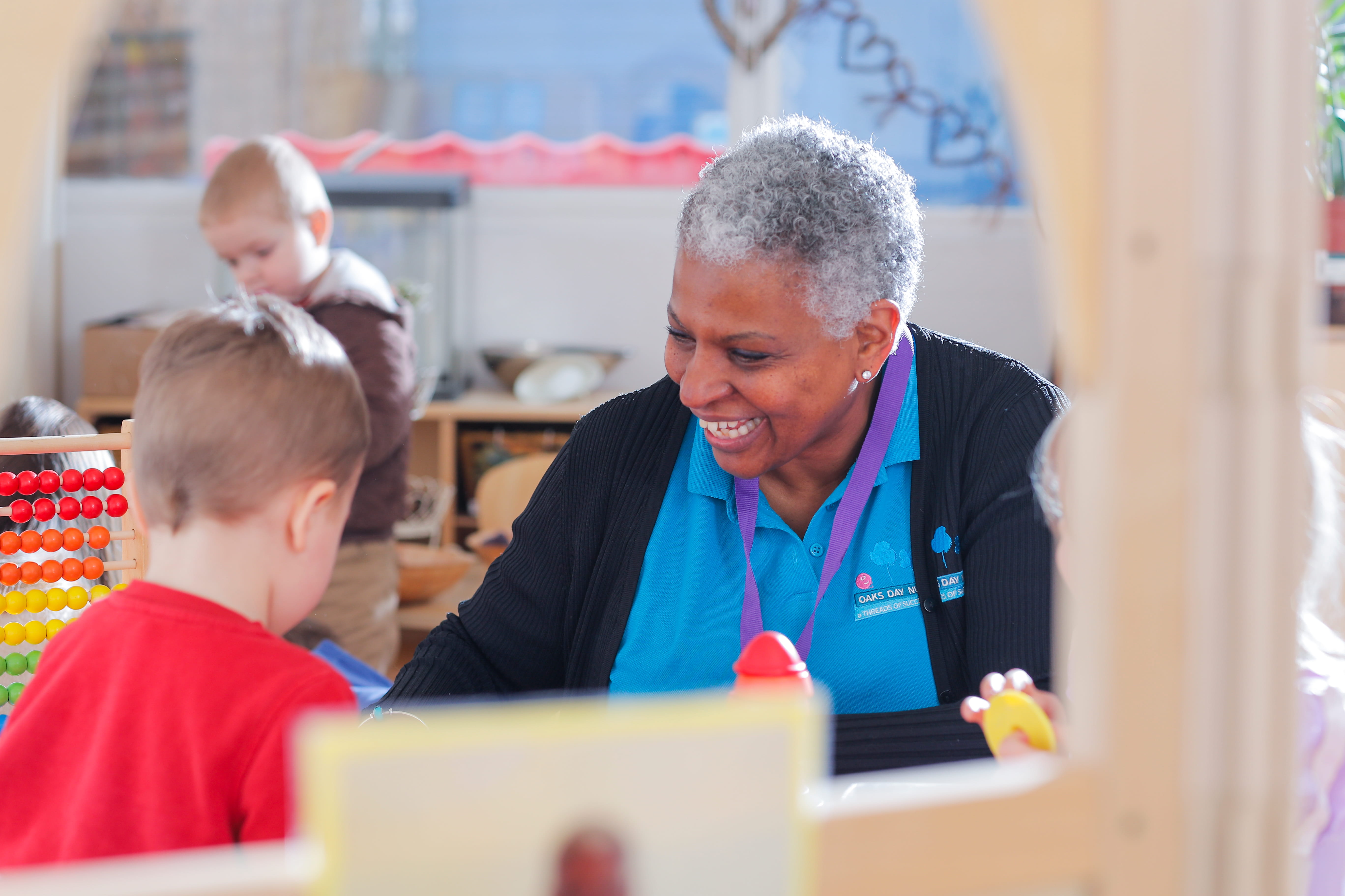 ALT - A smiling woman engages with young children in a classroom. She wears a blue shirt and purple lanyard. Bright toys and a wooden abacus are visible.