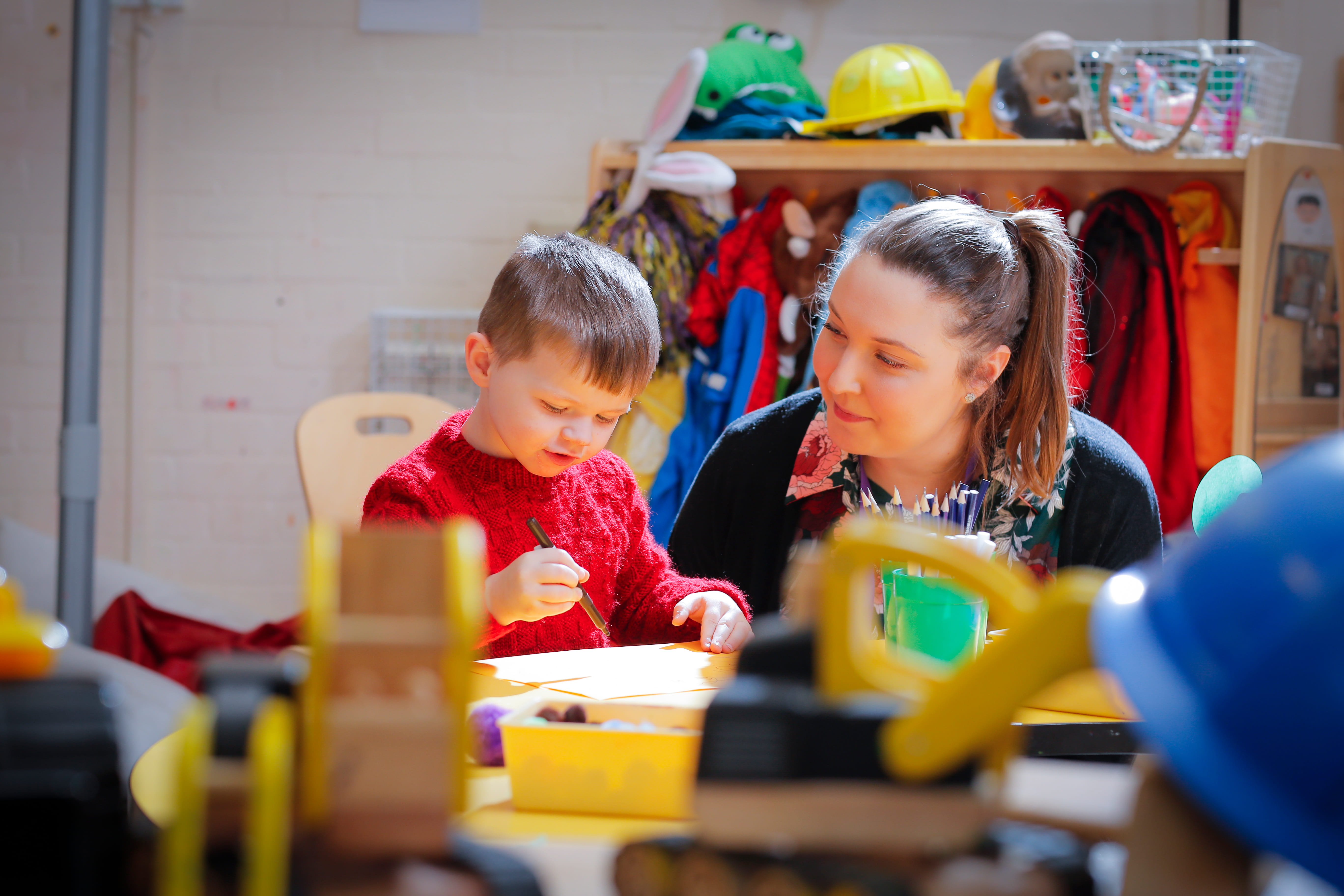 ALT - A child in a red sweater writes with a black pen, supervised by a smiling adult in a colourful room filled with toys and dress-up clothes