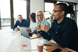 ALT - Four adults sitting at a desk one working on a laptop, smiling and talking to each other