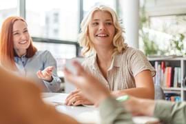 ALT - Two ladies sitting at a desk having a conversation, one lady is using her hands to express herself and smiling