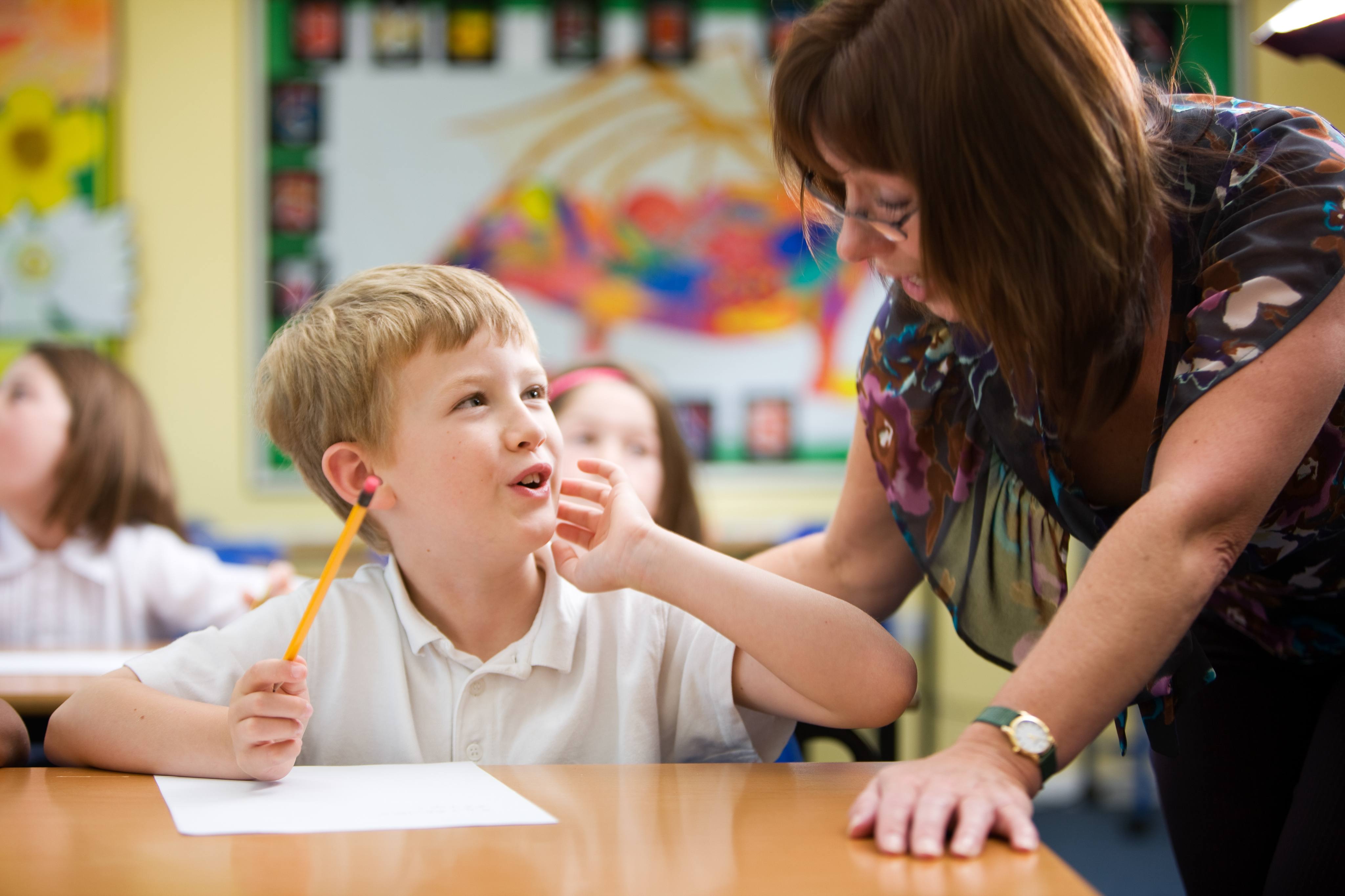 Alt - Child sitting at desk with teacher standing next to him talking to each other