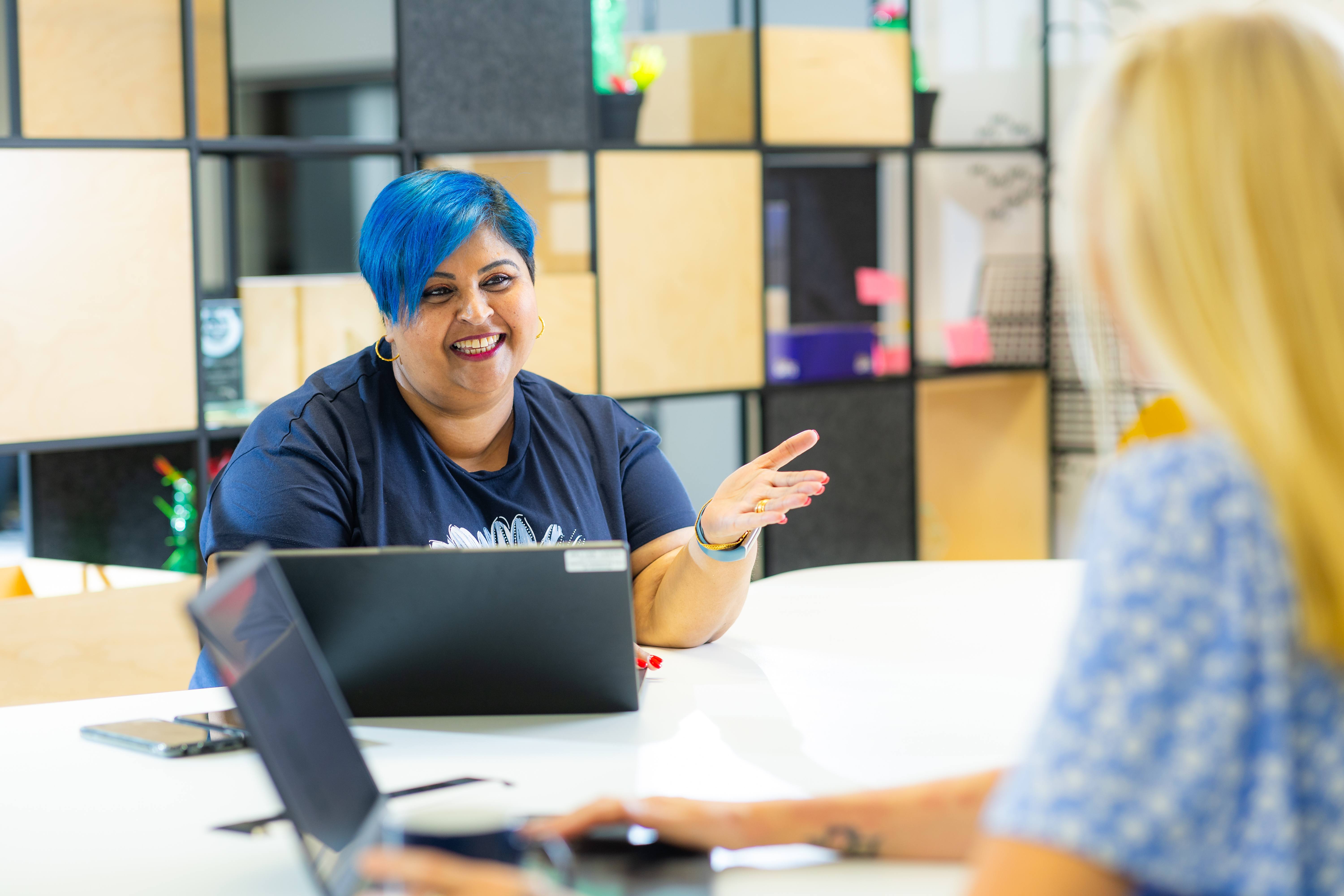 ALT - Two ladies sitting at a desk working on laptops talking to each other
