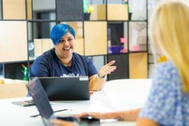 ALT - Two ladies sitting at a desk working on laptops talking to each other