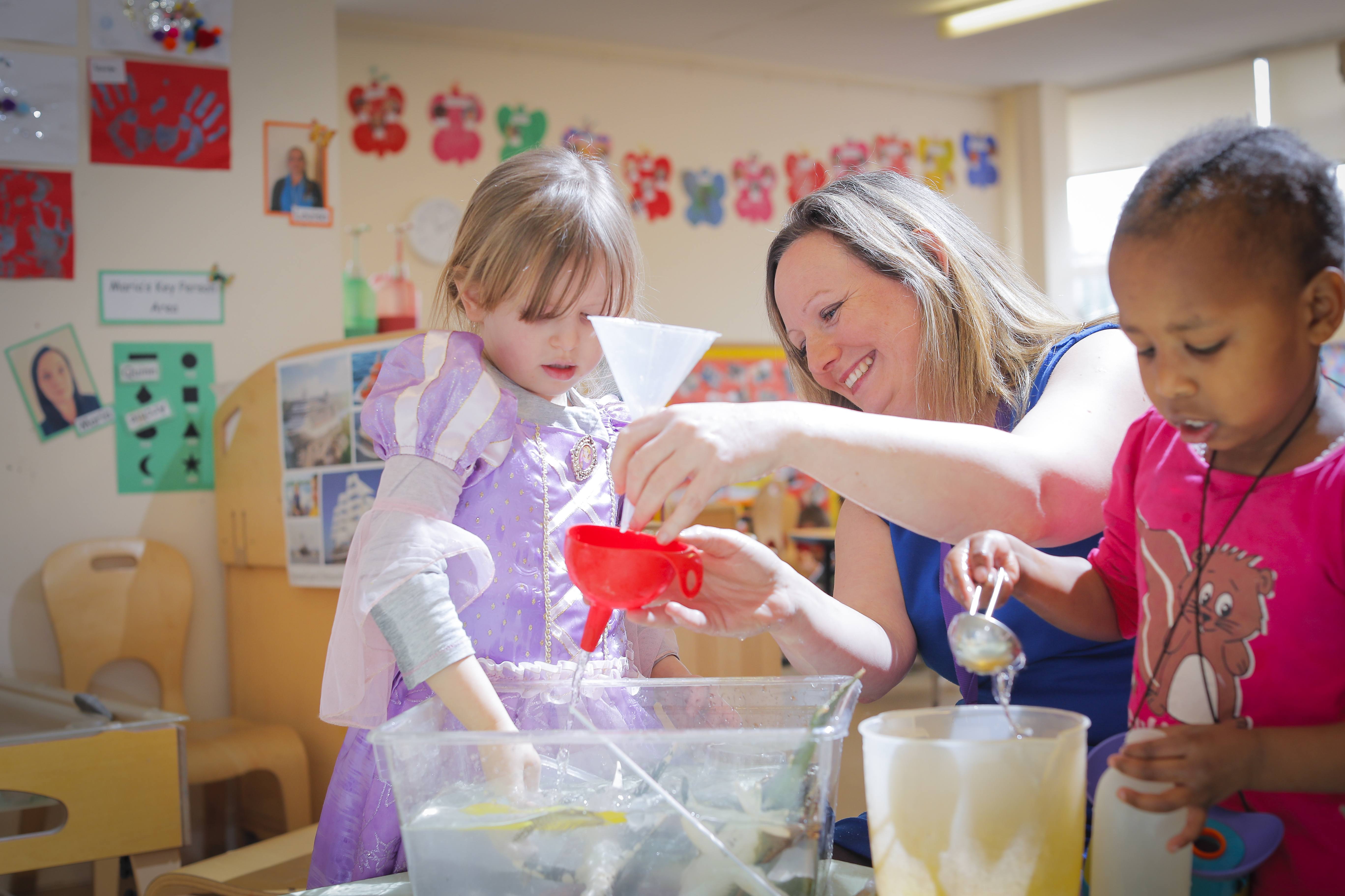 ALT - nursery teacher holding a funnel tipping water through it whilst a child puts her hand in the water to feel it
