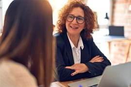 ALT - Two ladies talking to each other whilst sitting at a desk working on laptops