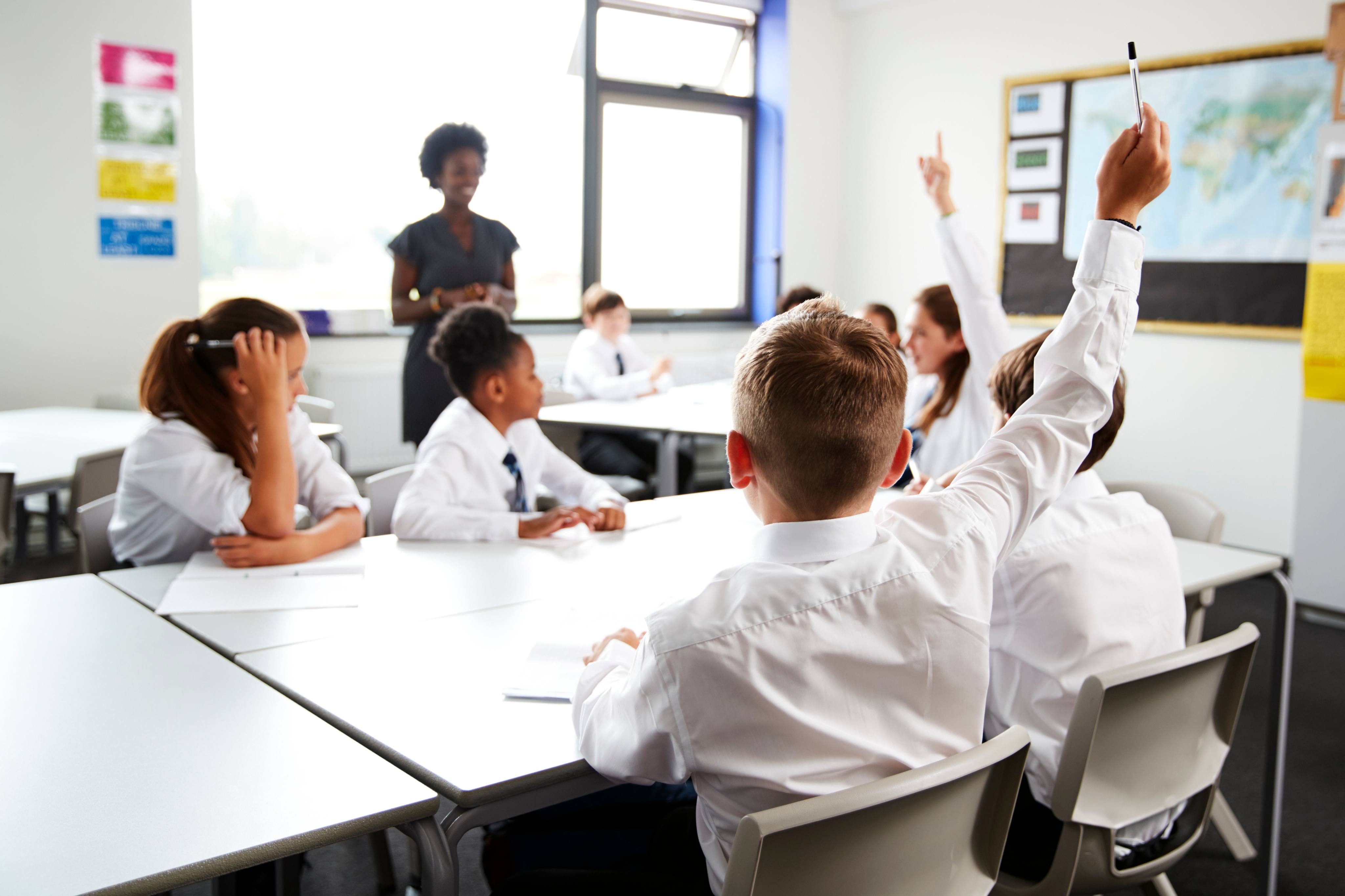 ALT - Children sitting and listening to teacher in the classroom, holding their hands up to answer the questions