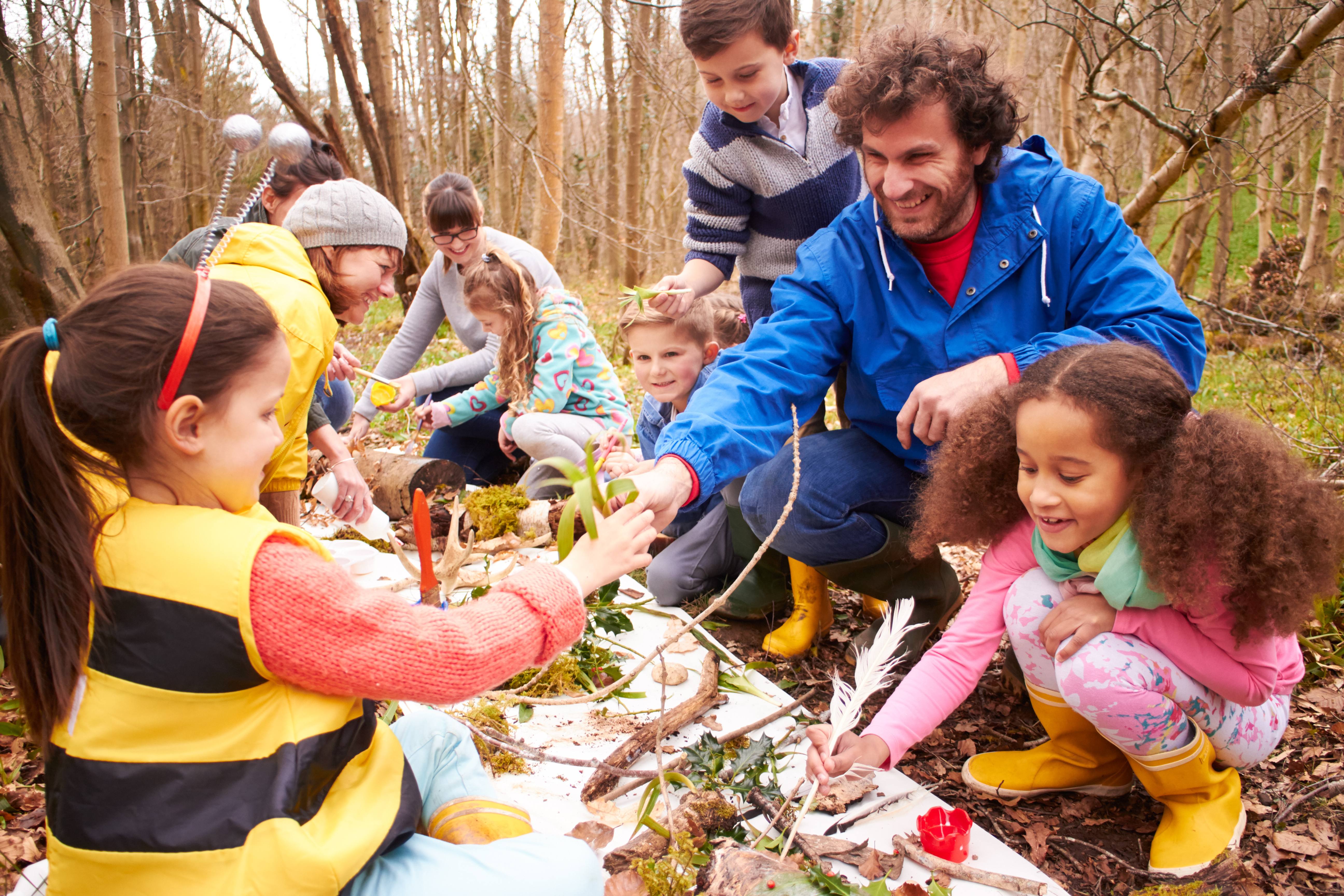 ALT - a group of young children and teachers sitting on the ground in the woods exploring leaves and twigs