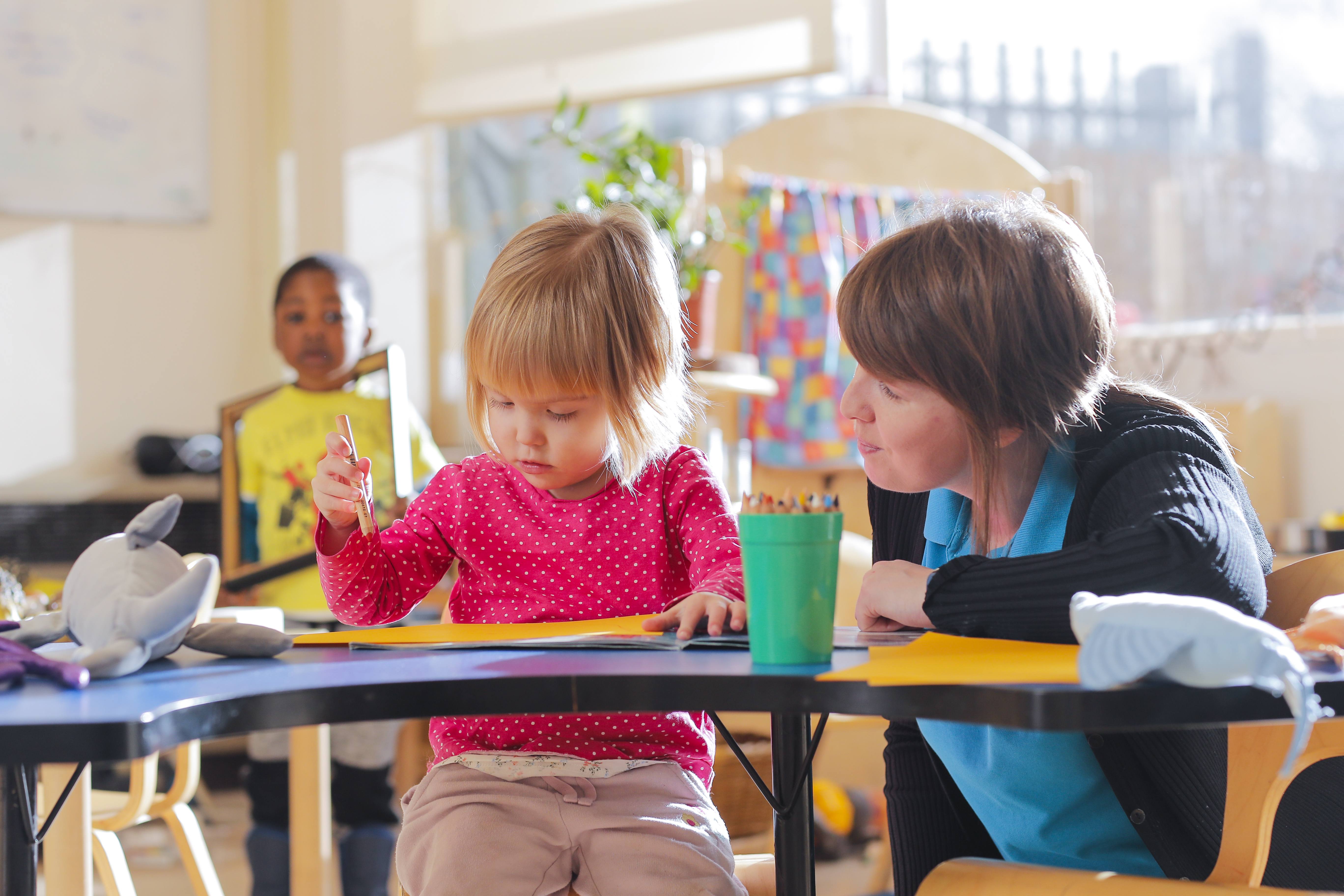 ALT - Nursery teacher sitting next to a child whilst she draws with a pencil