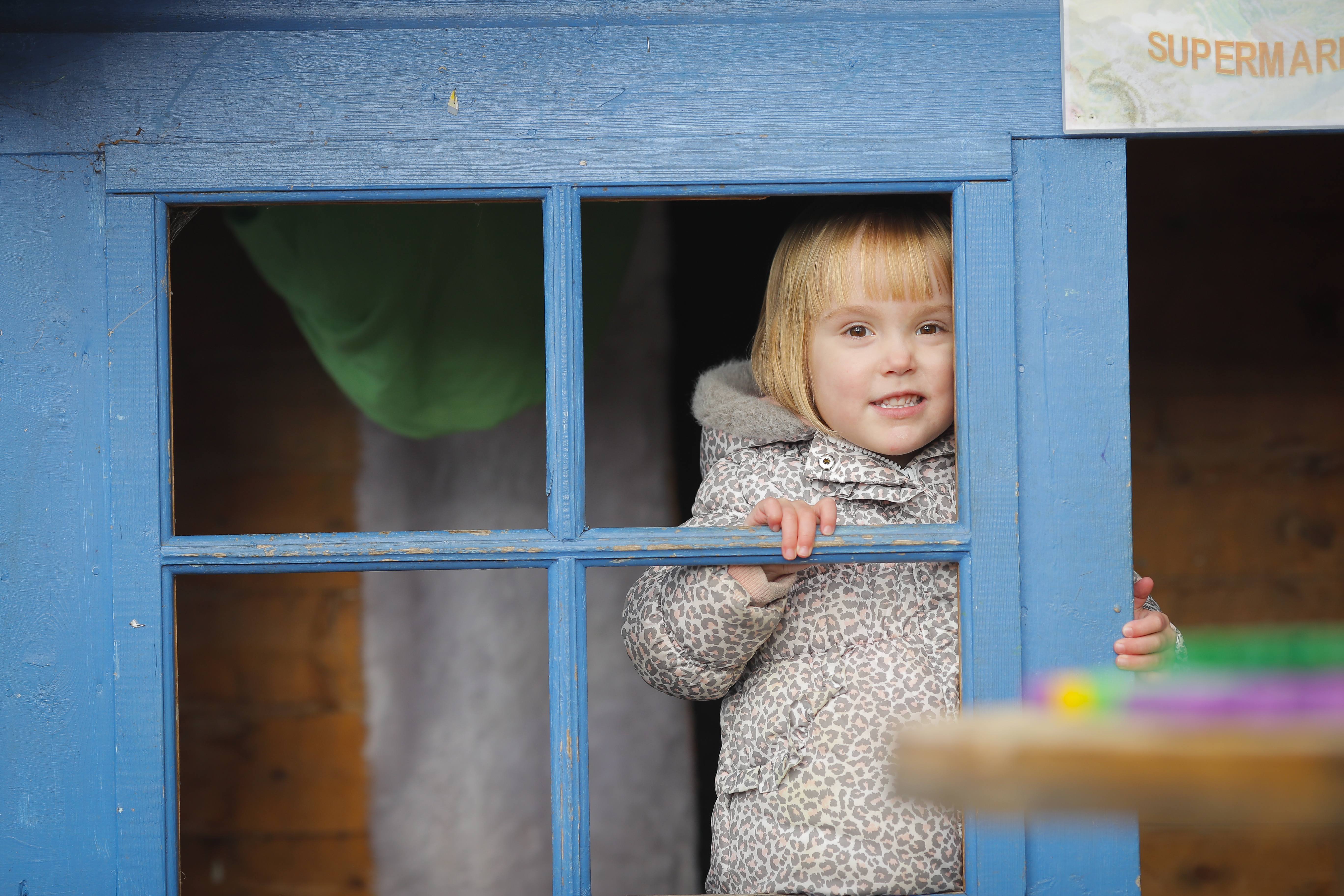 ALT - Child standing inside a wendy house smiling out of the window