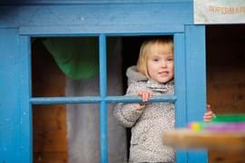 ALT - Child standing inside a wendy house smiling out of the window