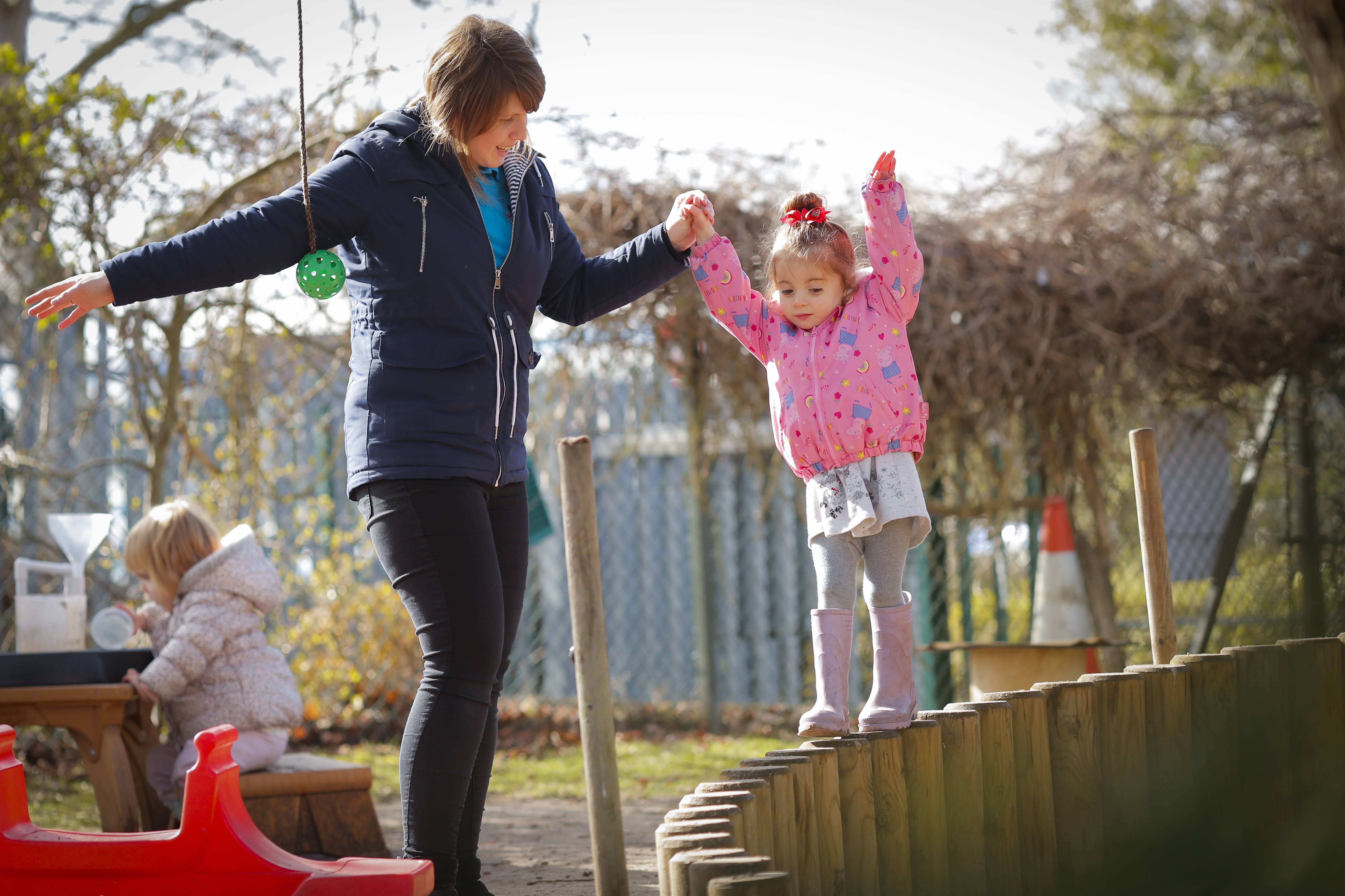 ALT - Nursery teacher holding a childs hand whilst she balances on a wall made of wood