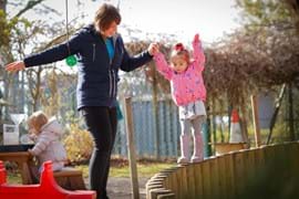 ALT - Nursery teacher holding a childs hand whilst she balances on a wall made of wood
