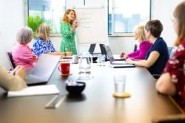 ALT - Adults sitting around a large desk working on laptops listening to trainer talk