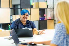 ALT - Two ladies sitting at desk with laptops in front of them smiling and talking