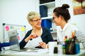 ALT - Two ladies sitting at a desk with a laptop having a conversation