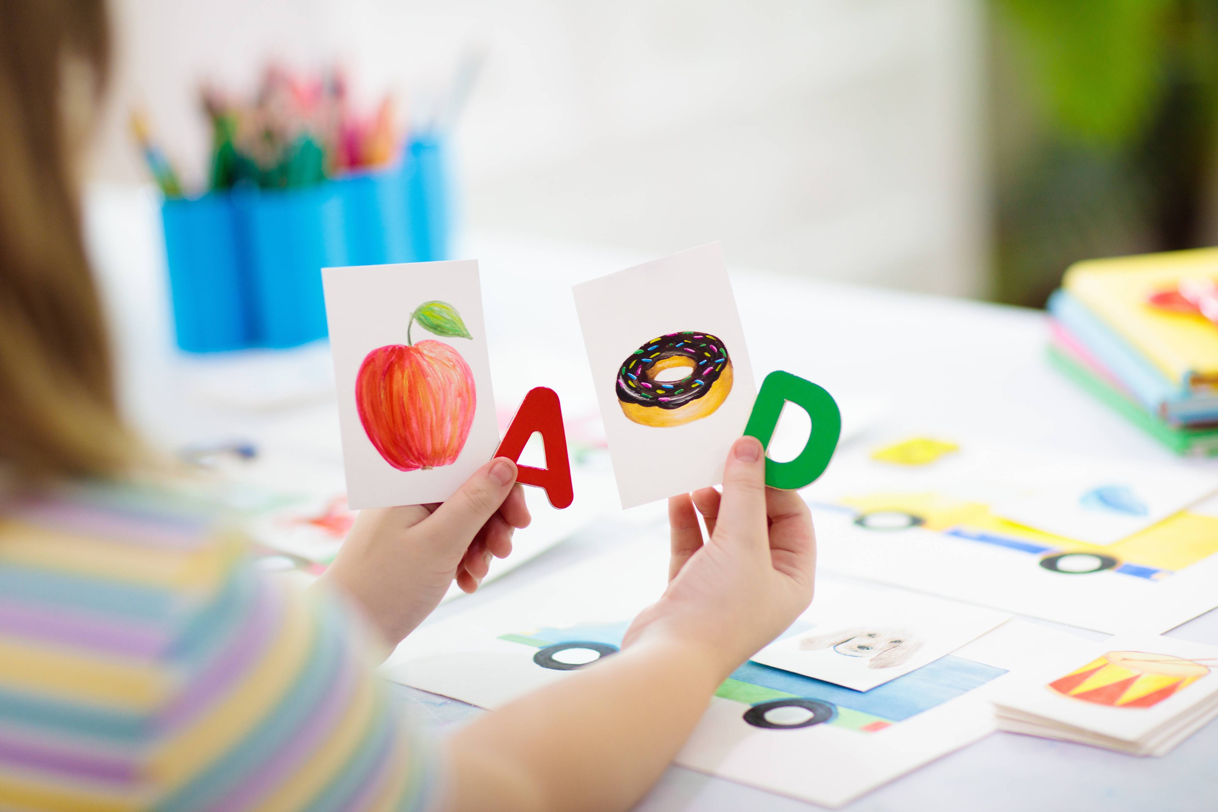 ALT - Child sitting at table holding the letter A with a picture for apple and D with a picture for donut in her hand