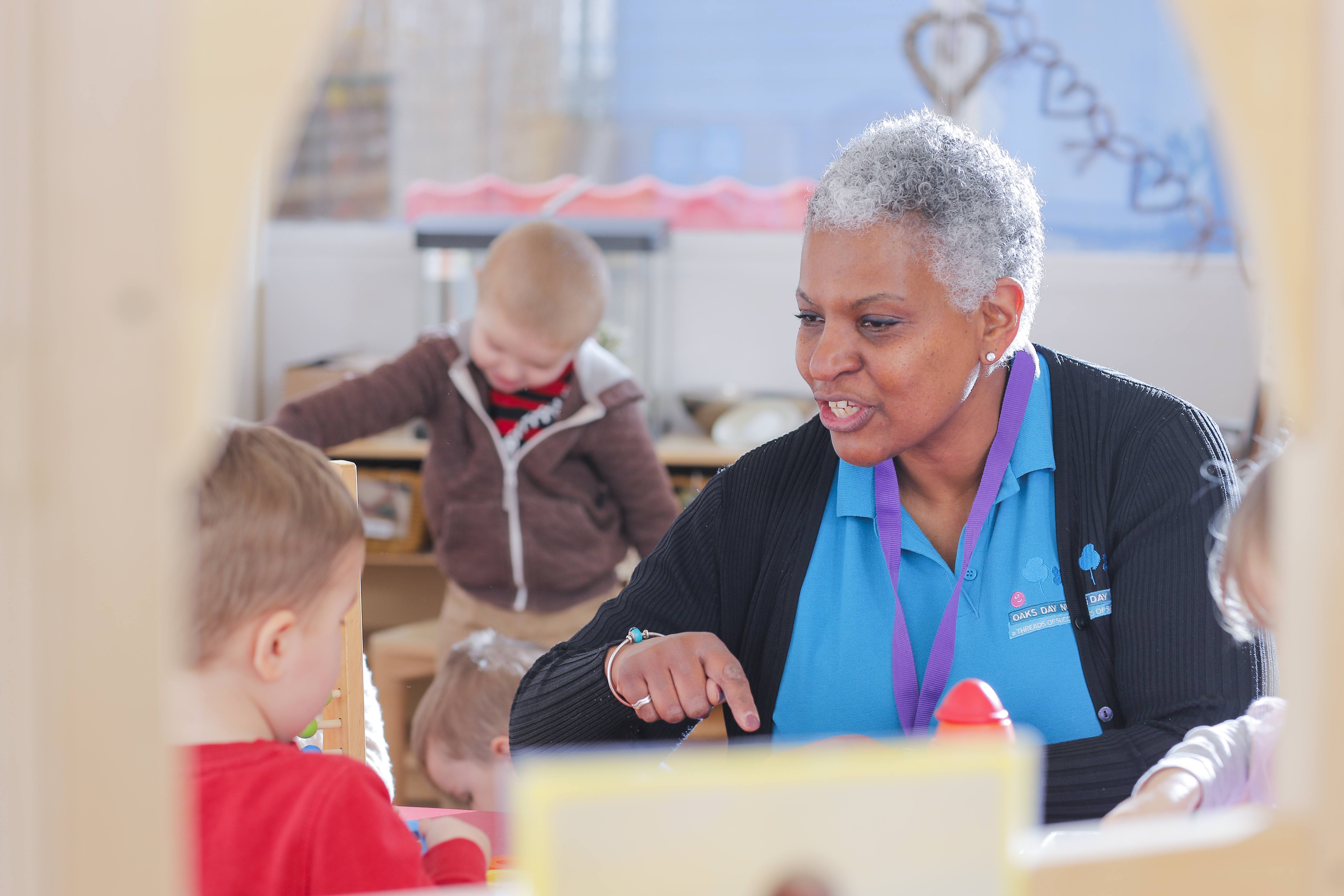 ALT - Nursery teacher talking to child sitting at a table pointing at a toy