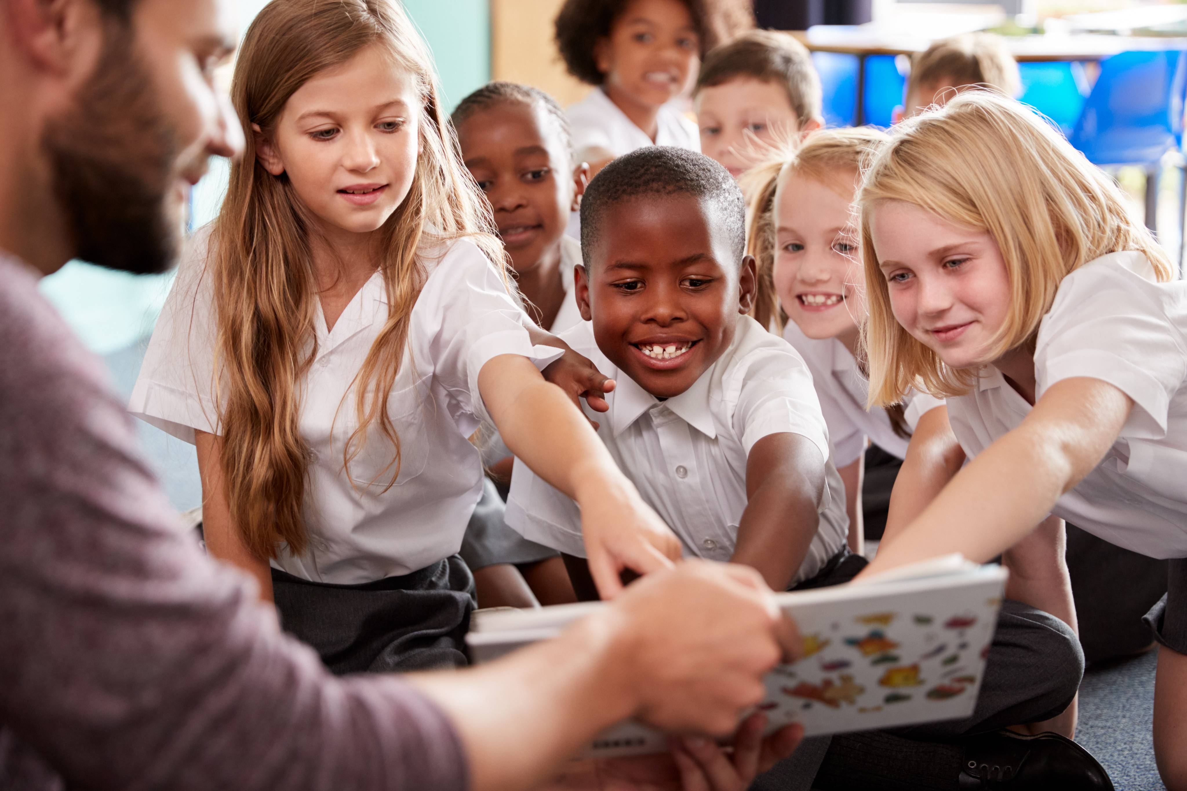 ALT - Teacher reading a book to a group of children, children are pointing and smiling at the pictures in the book