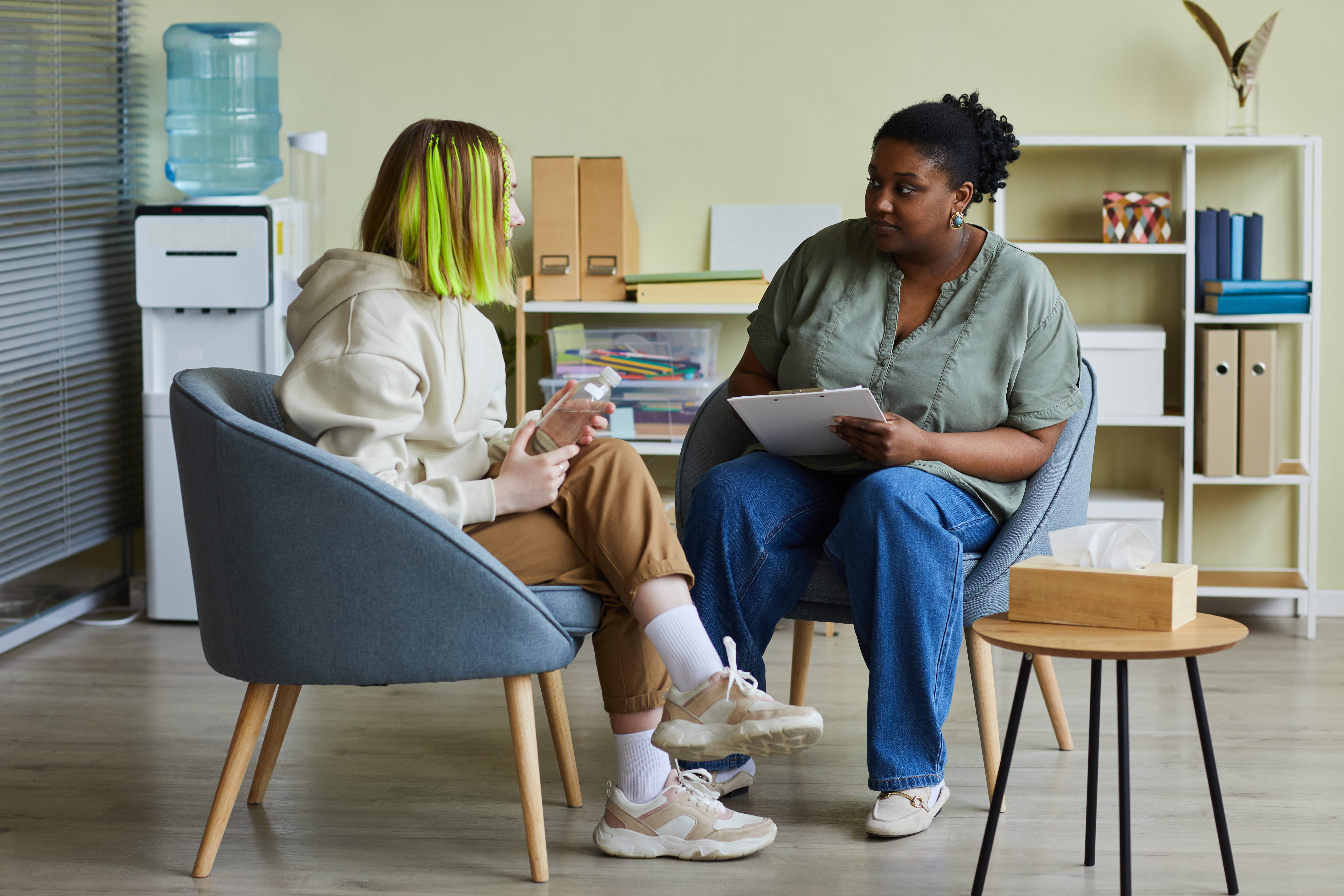 Two people sit in chairs facing each other in a cosy room with bookshelves.