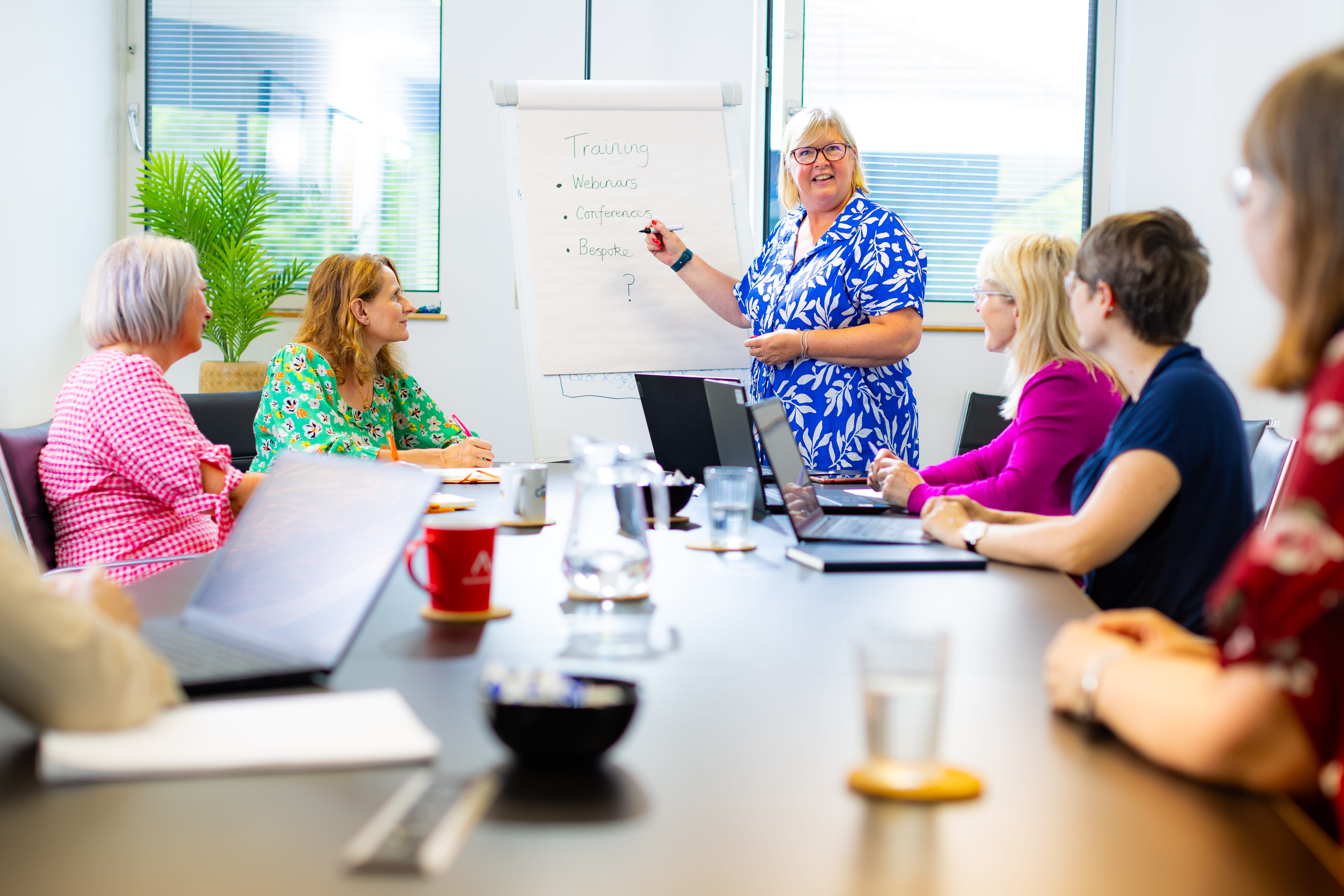 A woman in a blue floral dress presents a training topic on a flip chart in a bright conference room
