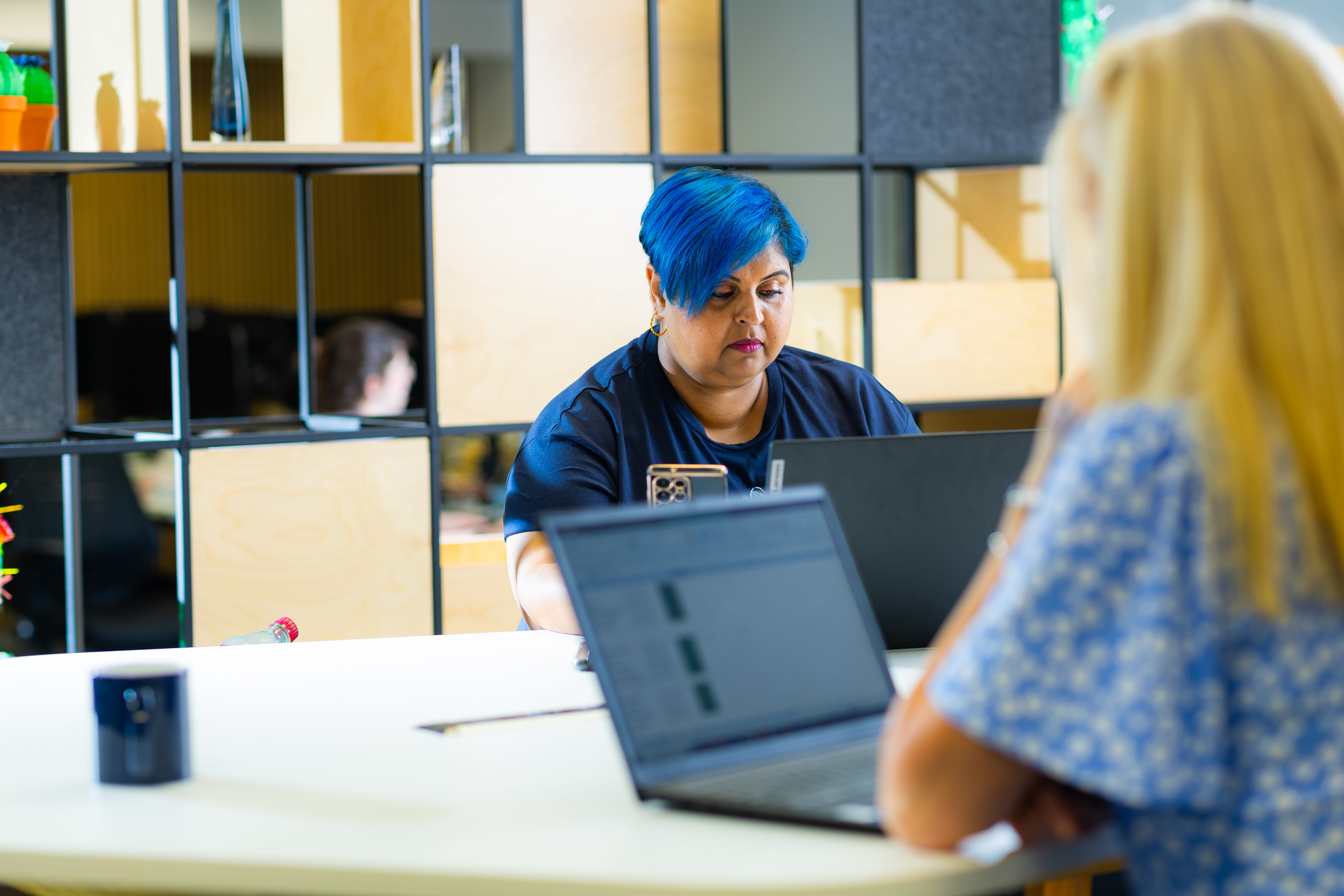 Two people working at laptops in a bright, modern office. One person with blue hair appears focused; the other, blurred in the foreground.