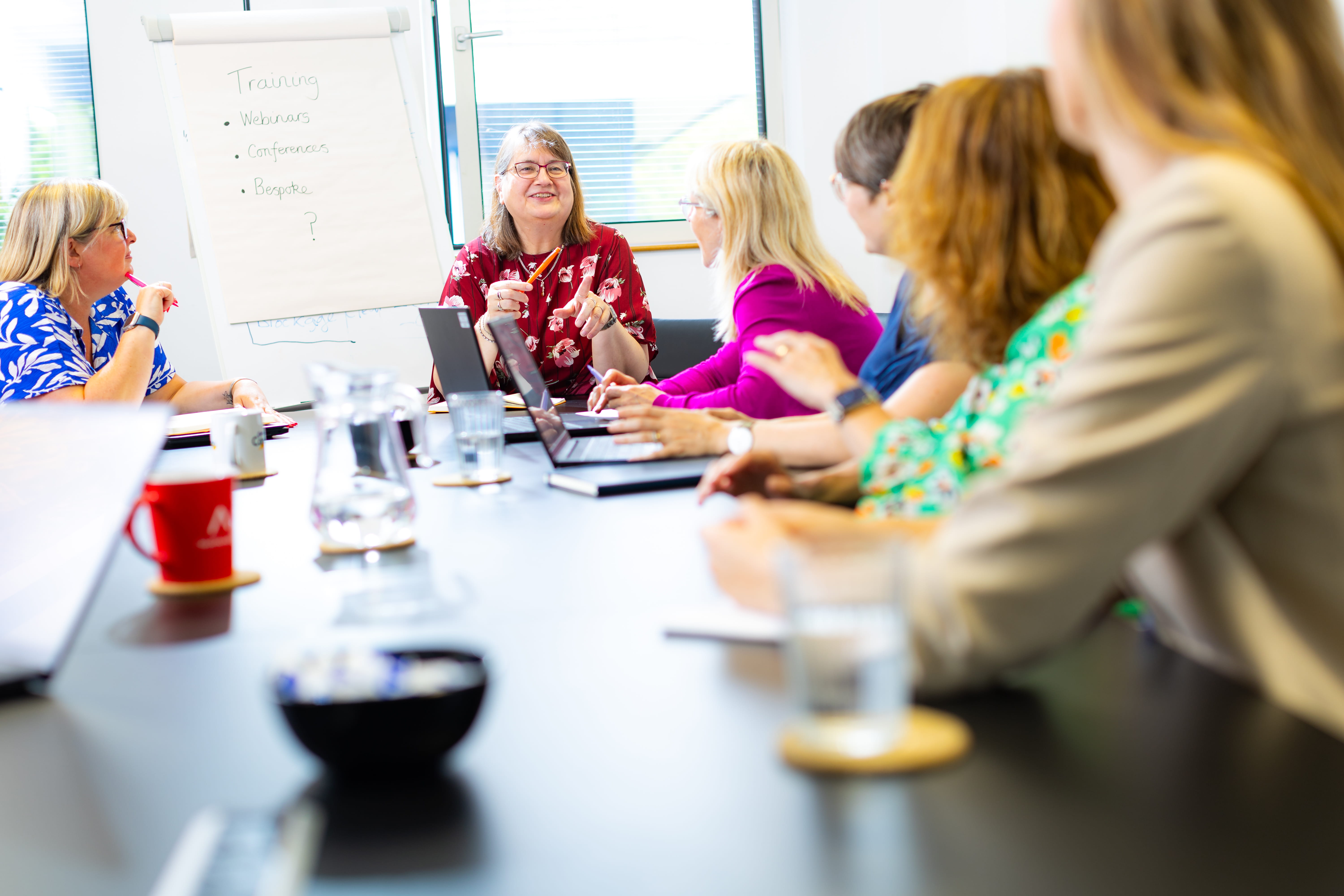 A diverse group of women sit at a conference table in a bright room, engaged in discussion