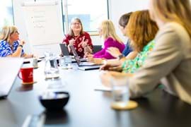 A diverse group of women sit at a conference table in a bright room, engaged in discussion